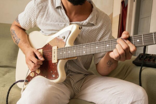 A close-up shot of a man sitting on a light green sofa playing an off-white electric guitar with a tortoise-shell pickguard. The man wears a white and black vertically striped short-sleeved button-up shirt and light-colored trousers, with visible tattoos on his arms. The shot focuses on his hands as he strums and frets the instrument, set against a neutral, airy indoor background.