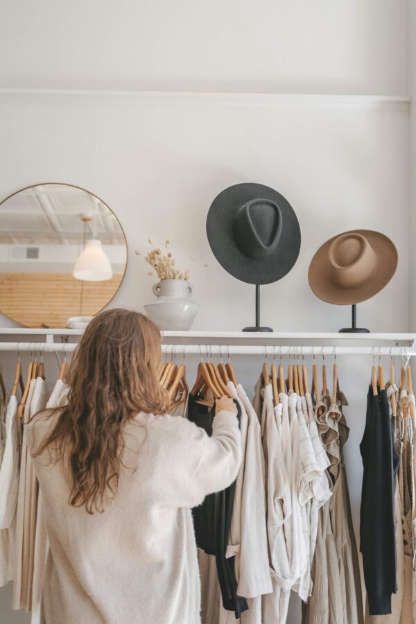 A rear view of a woman with long wavy hair browsing through a white clothing rack filled with neutral-toned garments on wooden hangers. Above the rack, a shelf displays a ceramic vase with dried flowers and two wide-brimmed fedora hats on black stands. To the left, a large circular mirror with a gold frame reflects a modern white pendant light and a warm wood-slat ceiling, creating a bright and airy retail aesthetic.