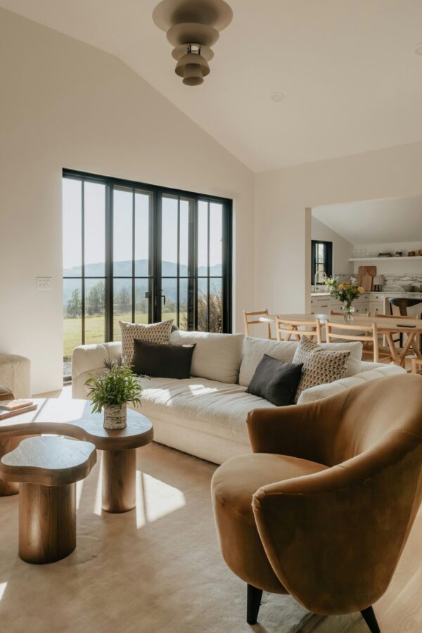 A sun-drenched, modern living room with organic textures. A cream-colored bouclé sofa is styled with dark brown and patterned throw pillows, sitting alongside a velvet cognac-colored armchair. In the foreground, a curved, solid-wood nesting coffee table holds a small potted plant. Large black-framed glass doors in the background reveal a bright, grassy landscape, while a light wood dining set is visible in the adjacent open-concept kitchen space.