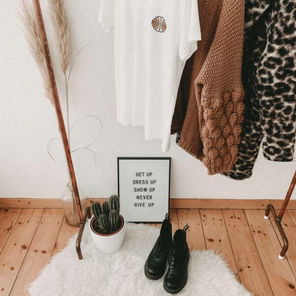 A stylish clothing vignette featuring an industrial-style rack made of copper piping. Hanging on the rack are a white t-shirt with a small graphic, a chunky brown knit cardigan, and a leopard print coat. Below, on a white faux-fur rug over light wood floors, sit black leather boots, a potted cactus, and a letter board that reads, 