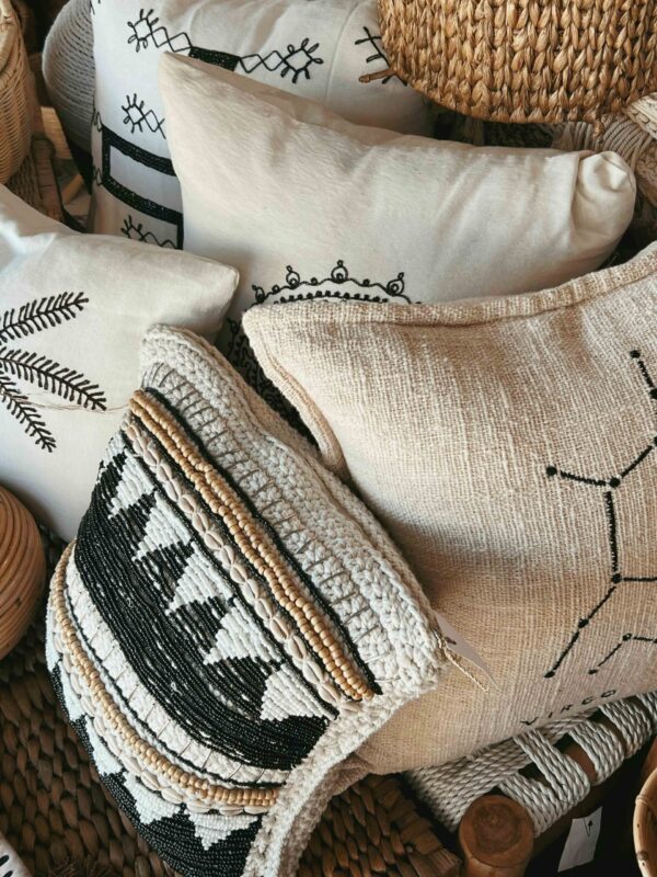 A close-up, high-angle shot of a pile of bohemian-style decorative pillows and woven baskets. The pillows feature various neutral-toned fabrics, including cream canvas with black embroidered tribal patterns, a burlap-textured cushion with a simple line drawing, and a highly detailed hand-knotted pillow accented with black beads, white shells, and tan wooden beads. The items are arranged in a textured, rustic display that emphasizes organic materials and intricate craftsmanship.