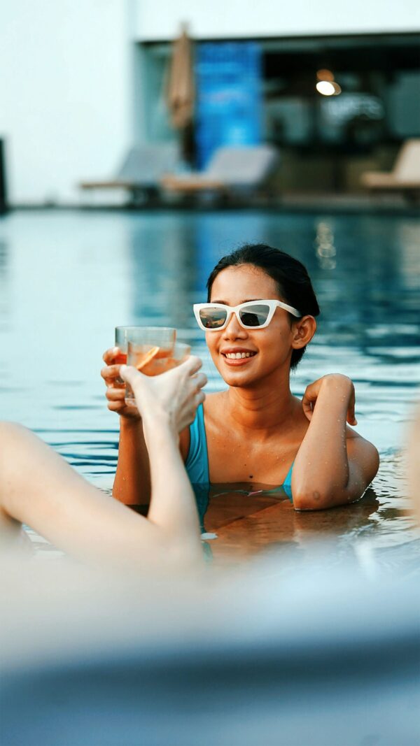 A young woman with a bright smile, wearing a turquoise swimsuit and white-framed cat-eye sunglasses, relaxes in a blue swimming pool. She is clinking glasses with a friend for a toast, holding a cocktail garnished with an orange slice. The background features a soft-focus resort setting with lounge chairs, all captured in vibrant, warm natural daylight.