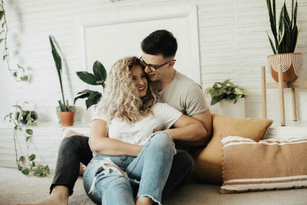 A young couple sitting playfully on a carpeted floor in a bright, modern living room. The woman, with long blonde curly hair, laughs as the man in glasses leans in close. They are surrounded by various indoor plants in terracotta and patterned pots, with mustard-yellow and striped throw pillows leaning against a white textured-brick wall. The scene is filled with soft, airy natural light, creating a warm and intimate home atmosphere.