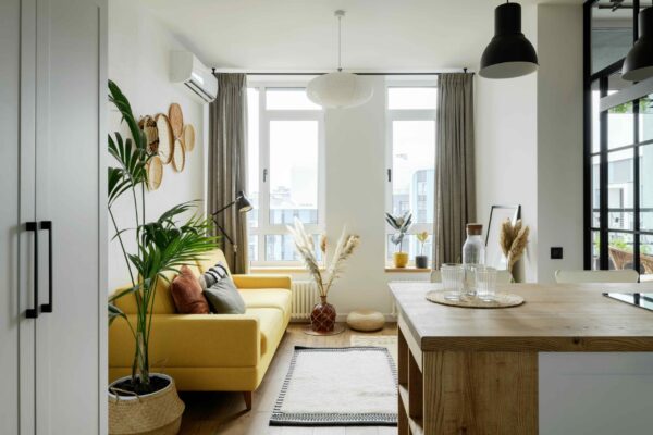 A bright, open-concept studio apartment featuring a living area and a wooden kitchen island in the foreground. The living space includes a vibrant yellow sofa accented with rust and grey pillows, set against a white wall decorated with woven baskets. Three distinct lighting sources are visible: a large white saucer-shaped pendant light over the center, a black industrial-style floor lamp behind the sofa, and a large black dome pendant light in the top right. Large windows with floor-to-ceiling grey curtains fill the space with natural light.