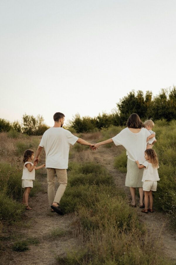 A rear view of a young family of five walking hand-in-hand down a narrow dirt path through a grassy field at golden hour. The parents and three young children are dressed in coordinated neutral and white linen clothing. The scene is bathed in soft, warm evening light with a hazy treeline in the background under a pale sky, evoking a sense of peaceful connection and natural beauty.