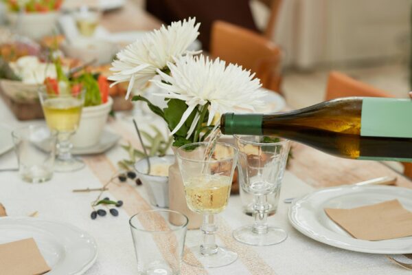A close-up of an elegant dinner table setting where white wine is being poured from a dark green bottle into a stemmed glass. The table is decorated with a white linen runner, large white chrysanthemum flowers in a rustic vase, and fresh olives on the branch. Soft, natural light illuminates the scene, highlighting the delicate glassware, white ceramic plates, and warm, inviting ambiance of a sophisticated gathering.