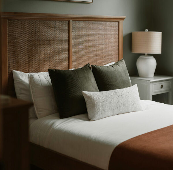 A close-up of a sophisticated bedroom featuring a high wooden headboard with a woven cane inset. The bed is layered with white linens, two dark olive-green velvet pillows, and a long white textured lumbar pillow. A white ceramic bedside lamp with a tan drum shade sits on a light grey nightstand, casting a soft glow against a muted sage-green wall. A rust-colored throw blanket is visible at the foot of the bed.