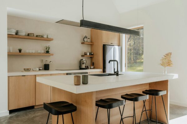 A modern, minimalist kitchen featuring light oak cabinetry and a large waterfall island with a thick white countertop. A long, linear black pendant light hangs horizontally above the island, which is paired with three black sculpted wooden barstools. The back wall includes open wooden shelving with artisanal ceramics and a built-in stainless steel refrigerator. Natural light streams in from a large window overlooking a wooded backyard, highlighting the clean lines and organic textures of the space.