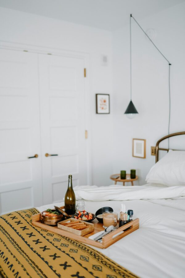 A bright, minimalist bedroom featuring a wooden breakfast tray on a bed with white linens and a yellow patterned throw. The tray holds a bottle of wine, fresh fruit, and coffee. A black cone-shaped pendant light hangs from a thin cable over the bedside, while the bed frame and door hardware feature elegant brass finishes. The room is decorated with small framed art pieces on a plain white wall, creating a clean, high-end residential aesthetic.