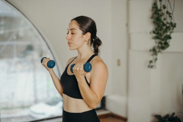 A young woman with her hair in a ponytail and wearing black athletic gear performs bicep curls with blue dumbbells in a minimalist indoor space. She has her eyes closed in a focused, meditative expression. In the background, a large arched window provides a view of a soft-focus urban setting, filling the clean, white room with soft, diffused natural light.