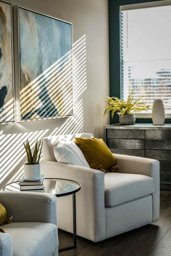 A sunlit modern living room corner featuring a plush, cream-colored armchair accented with a mustard-yellow velvet pillow. Stark, linear shadows from window blinds create a rhythmic pattern across a large abstract blue and tan canvas painting on the wall. A small glass side table holds a potted succulent, while a dark wood dresser with a green fern sits beneath a window in the background.